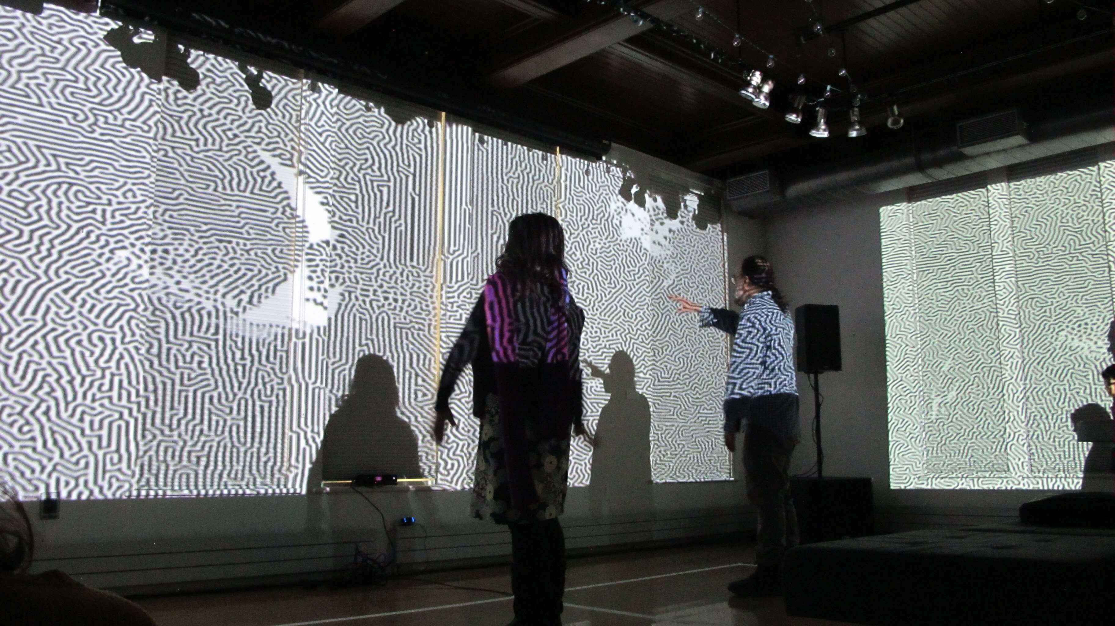 Image of people dancing in front of a large projection. The projection is a colorful abstract pattern that changes with the movement of the dancers. The dancers are silhouetted against the projection, creating a striking contrast.