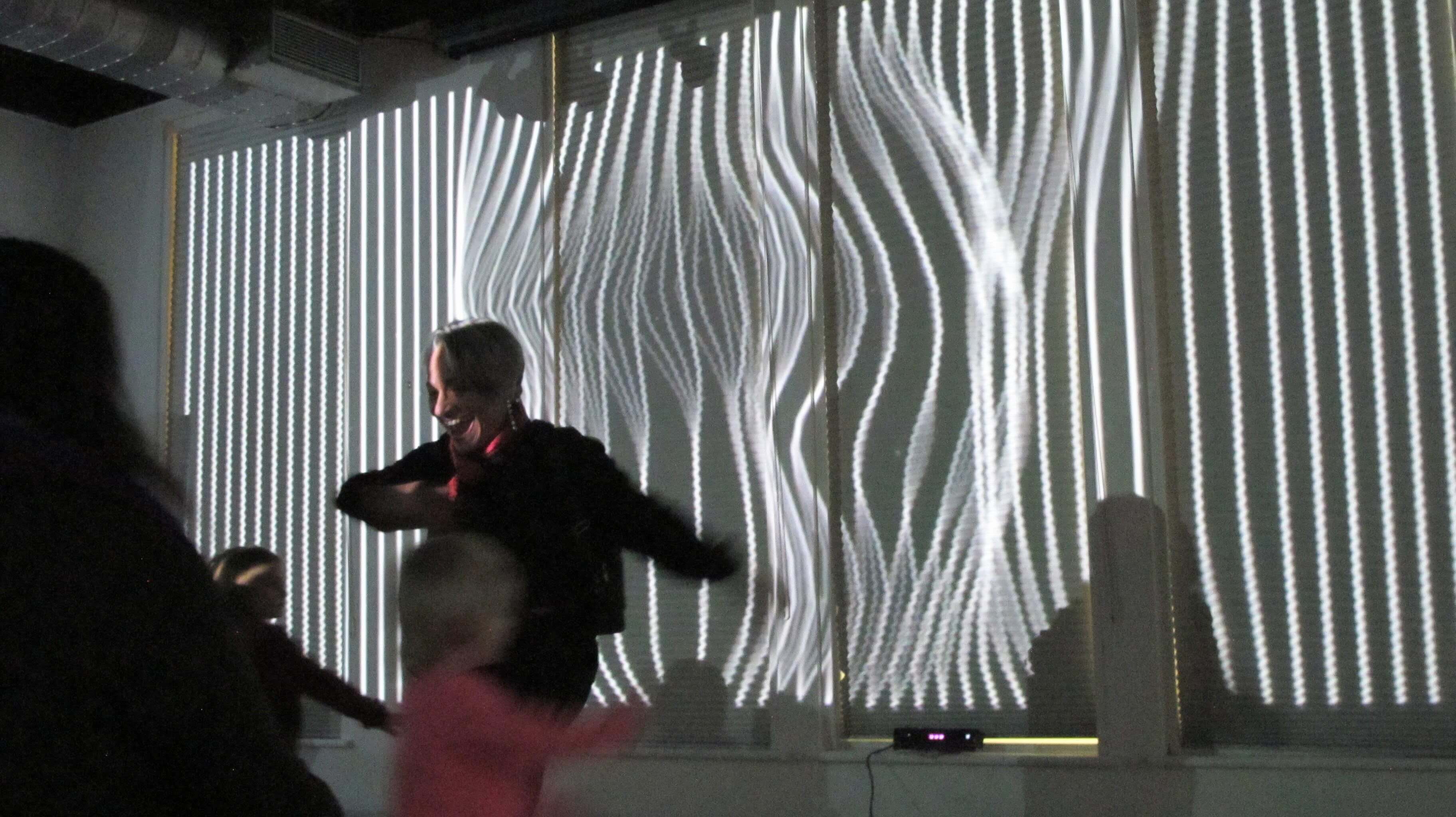 Image of people dancing in front of a large projection. The projection is a colorful abstract pattern that changes with the movement of the dancers. The dancers are silhouetted against the projection, creating a striking contrast.