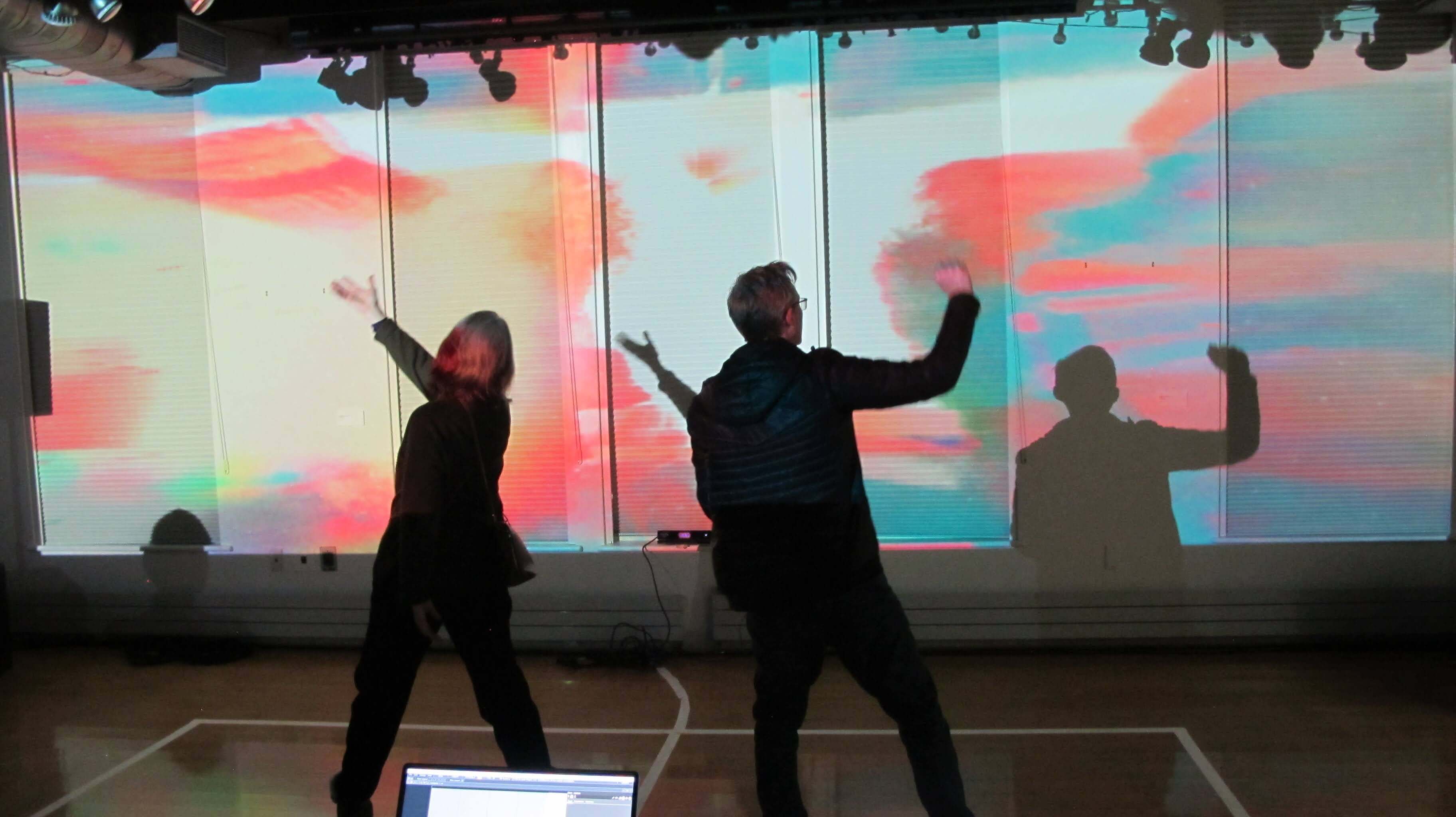Image of people dancing in front of a large projection. The projection is a colorful abstract pattern that changes with the movement of the dancers. The dancers are silhouetted against the projection, creating a striking contrast.
