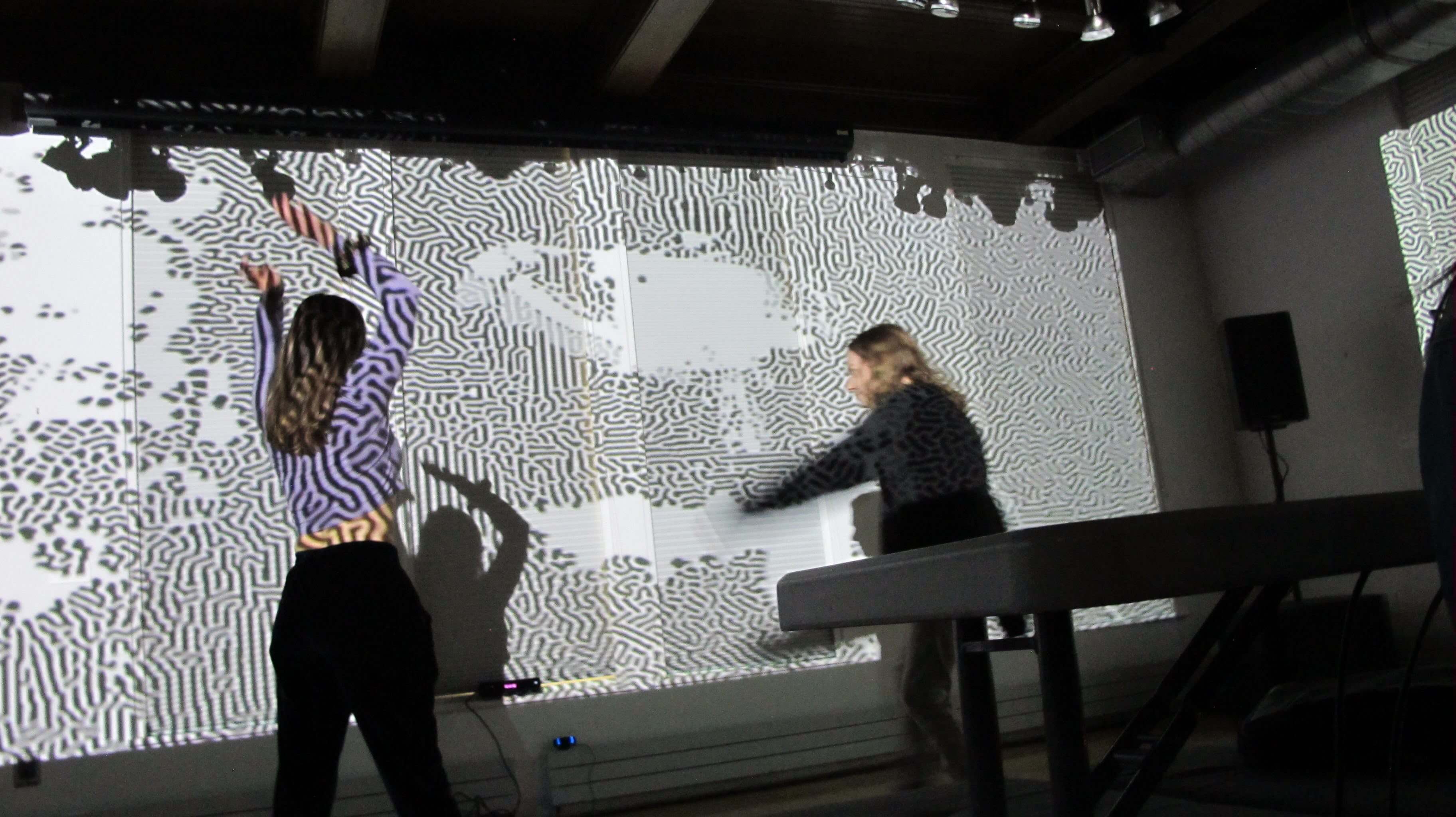 Image of people dancing in front of a large projection. The projection is a colorful abstract pattern that changes with the movement of the dancers. The dancers are silhouetted against the projection, creating a striking contrast.