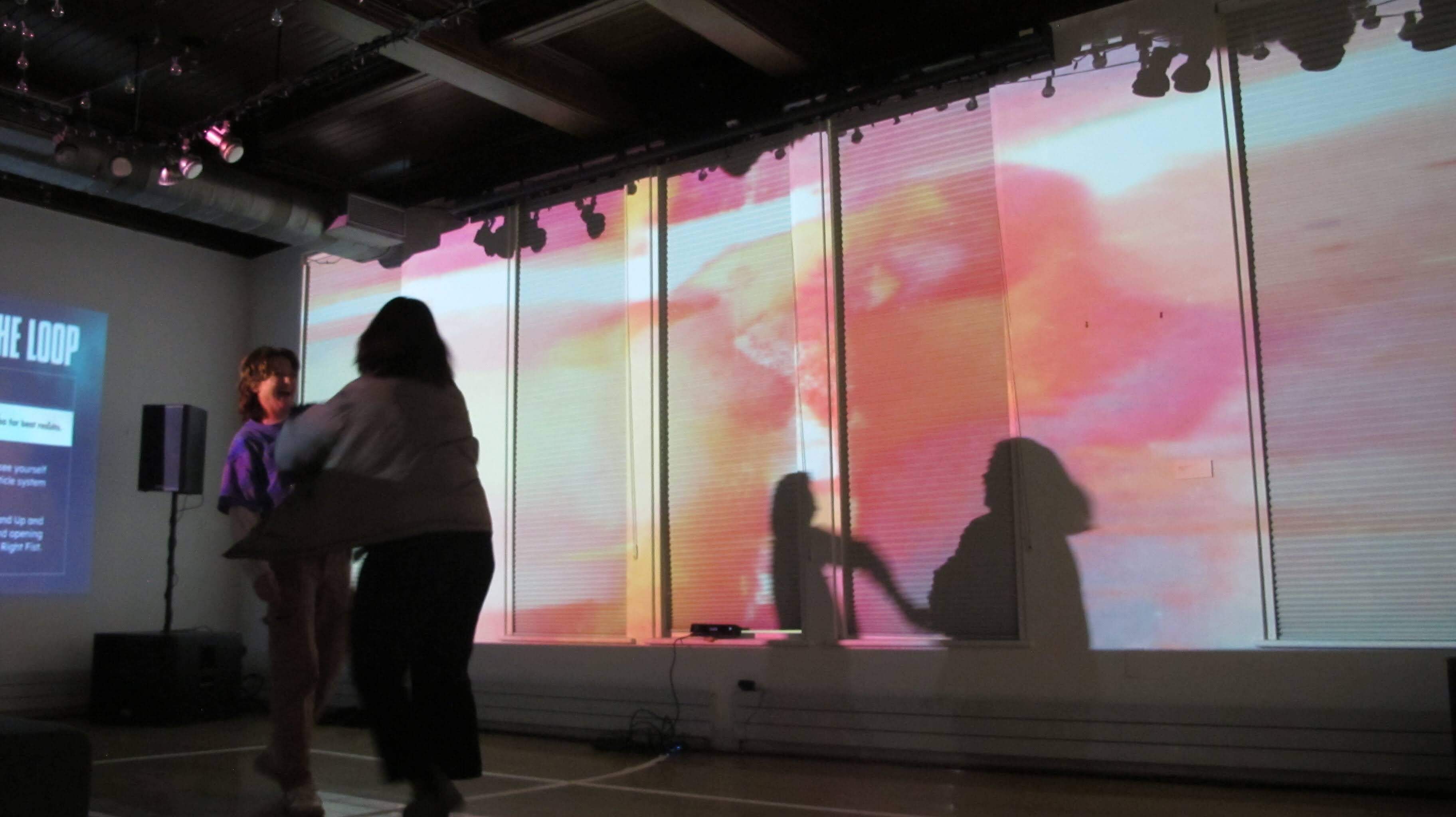 Image of people dancing in front of a large projection. The projection is a colorful abstract pattern that changes with the movement of the dancers. The dancers are silhouetted against the projection, creating a striking contrast.