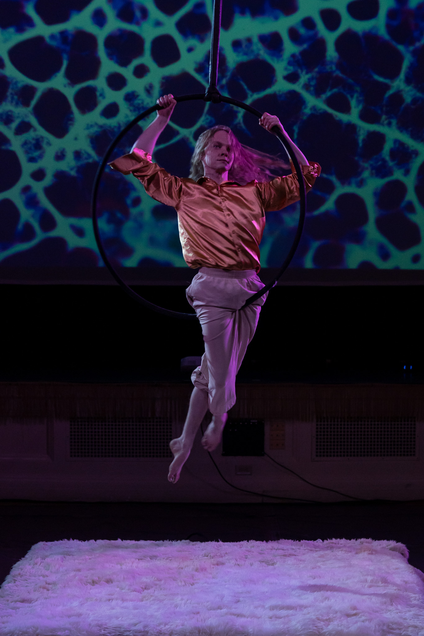A photograph of an aerialist performing in front of a large projection screen with colorful visuals.  The aerialist is silhouetted against the projection, creating a striking contrast.