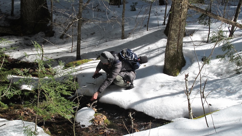 photo of ben recording water sounds in the field with a microphone and a portable recorder. The photo is taken in the winter, and there is snow on the ground. Ben is wearing a winter coat and a hat. He is standing next to a stream, and there are trees in the background.